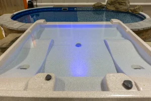 Modern hot tub with blue LED lighting and backyard pool visible behind it