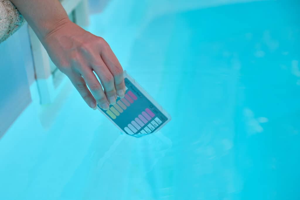 Hand dipping a pool water test strip beside its color chart in a clear backyard pool, the twice-a-week testing habit Cincinnati Pool and Patio recommends for Greater Cincinnati pool owners