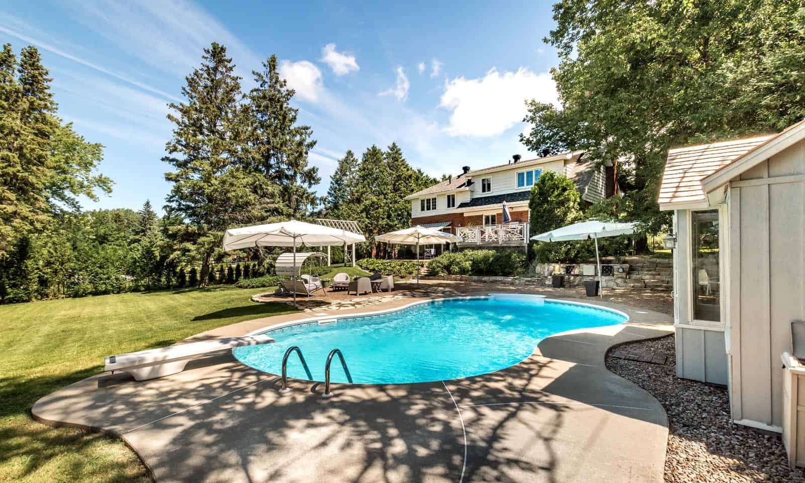 Kidney shaped inground vinyl liner pool with a concrete deck, a diving board and an equipment shed behind a two story brick home, the kind of backyard pool a heater keeps comfortable into fall